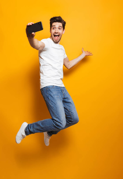 Full length portrait of happy man with brown hair jumping and screaming while taking selfie on black smartphone isolated over yellow background - Foto, Imagem