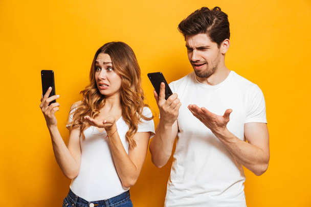 Confused young couple looking at mobile phones isolated over yellow background - Φωτογραφία, εικόνα
