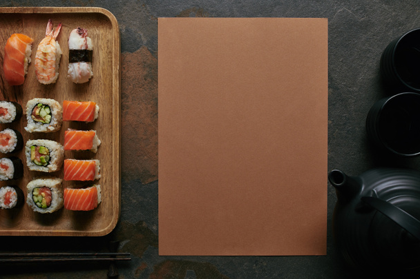 vlakke leggen met sushi ingesteld op houten plaat, zwarte theepot en kopjes op donkere tafelblad - Foto, afbeelding