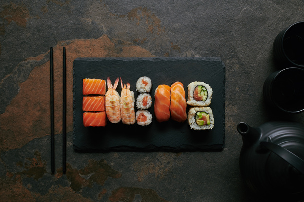 pohled shora sushi set na černé břidlicové desky, konvice a šálky čaje na tmavý podklad - Fotografie, Obrázek