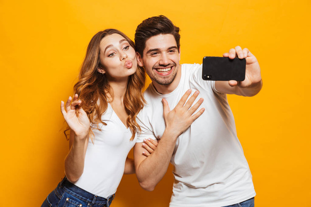 Image of beautiful content man and woman smiling and taking selfie photo on smartphone isolated over yellow background - 写真・画像