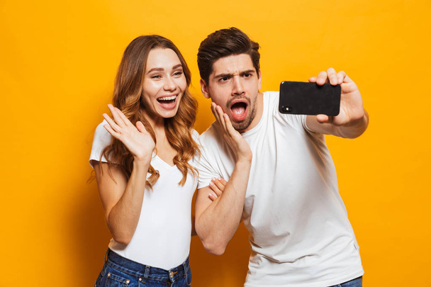 Portrait of happy man and woman smiling and taking selfie photo on smartphone isolated over yellow background - Foto, Bild