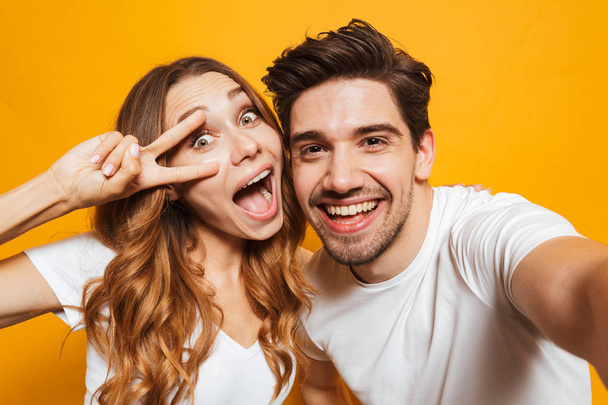 Portrait of attractive man and woman taking selfie photo and showing peace sign isolated over yellow background - Fotografie, Obrázek