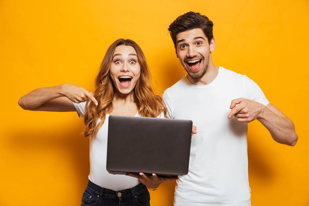 Portrait of pleased positive man and woman holding and pointing fingers at black laptop isolated over yellow background - Fotó, kép