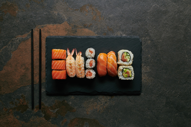pohled shora uspořádány sushi na černé břidlicové desky a hůlky na tmavý podklad - Fotografie, Obrázek