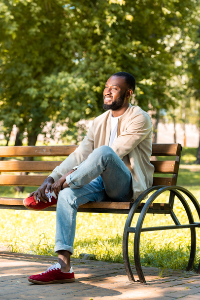 handsome smiling african american man sitting on wooden bench in park and looking away - Valokuva, kuva
