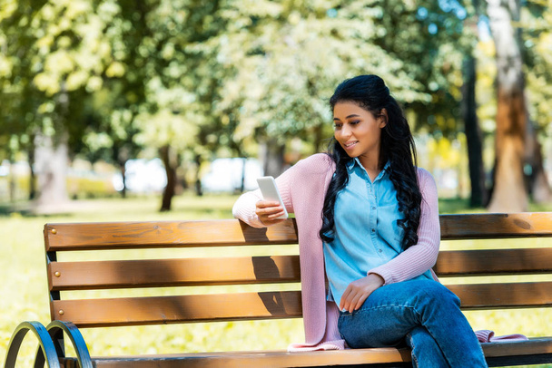 vidám vonzó afrikai-amerikai nő használ smartphone, a fa pad a parkban - Fotó, kép