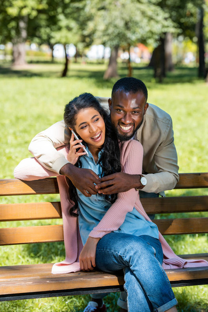 african american boyfriend hugging girlfriend while she talking by smartphone on bench in park - Photo, Image