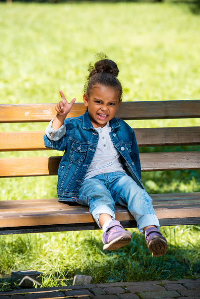 grimacing african american kid gesturing and sitting on wooden bench - Фото, изображение