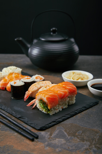 food composition with sushi set, ginger and soya sauce in bowls, teapot and cups with tea on dark surface - Valokuva, kuva