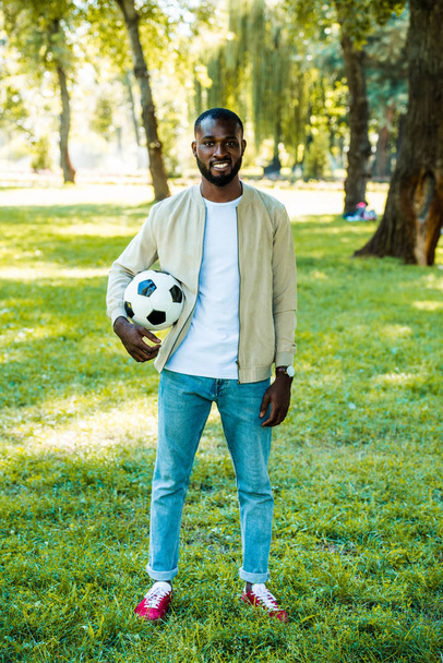 sorrindo bonito afro-americano homem de pé com bola de futebol no parque
 - Foto, Imagem