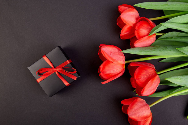 Scatola regalo nera con nastro rosso vicino bouquet tulipano rosso su sfondo nero. Piatto. Festa della mamma o della donna. Biglietto d'auguri. Ricevuto. Primavera
.  - Foto, immagini