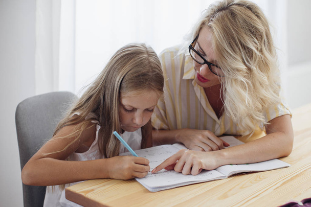 Cute blonde girl writing in her notebook with her mother helping. - Foto, immagini