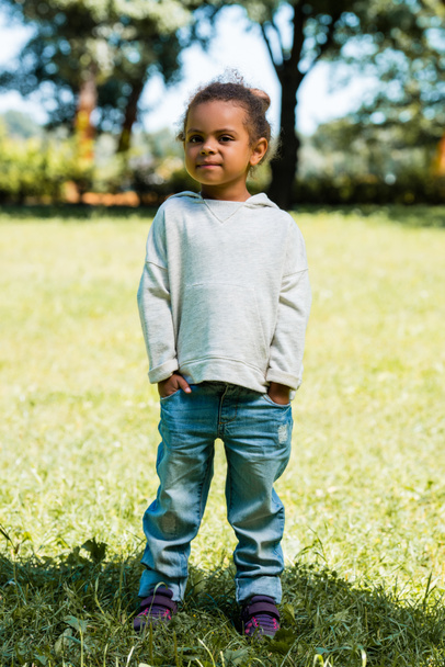 adorable african american child in jeans and sweater standing in park and looking at camera - Фото, зображення