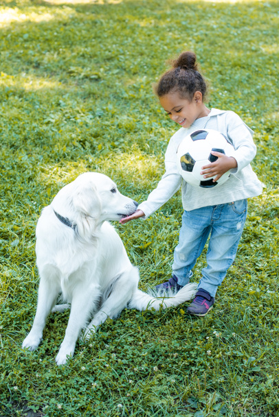 αξιολάτρευτο παιδί αφρικανική αμερικανική palming σκυλί και κατοχή μπάλας ποδοσφαίρου στο πάρκο - Φωτογραφία, εικόνα