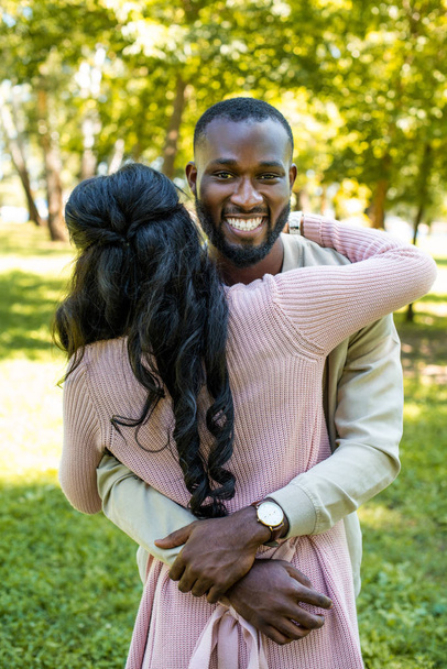 smiling african american boyfriend hugging girlfriend and looking at camera in park - 写真・画像