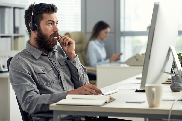 Brodaty mężczyzna w biurko do pracy w biurze przy użyciu komputera i mówiąc do klienta w zestaw słuchawkowy  - Zdjęcie, obraz