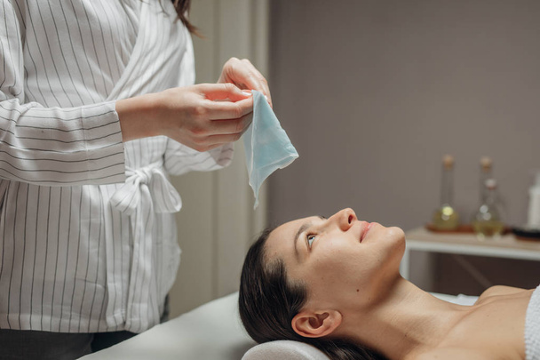 Mulher cosmetologista caucasiana colocando uma máscara facial no rosto de uma bela jovem branca deitada na cama de massagem
. - Foto, Imagem