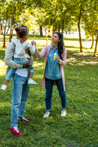 african american mother helping daughter blowing soap bubbles in park - 写真・画像
