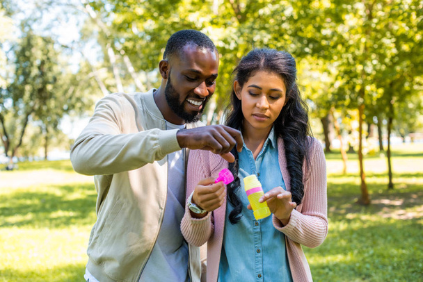 african american couple having fun with bottle for soap bubbles in park - Photo, Image