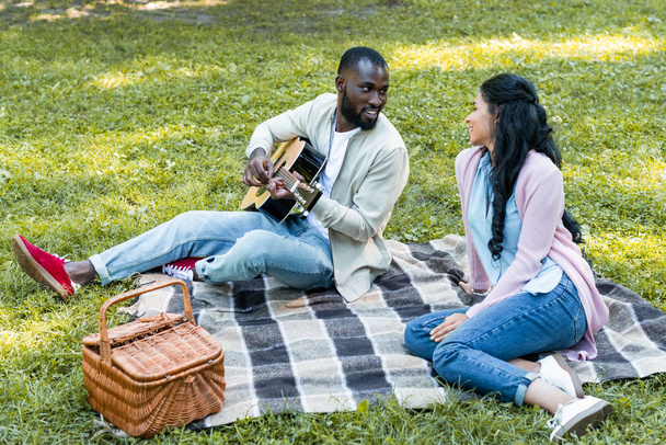 novio afroamericano tocando la guitarra acústica para su novia en el picnic en el parque
 - Foto, Imagen