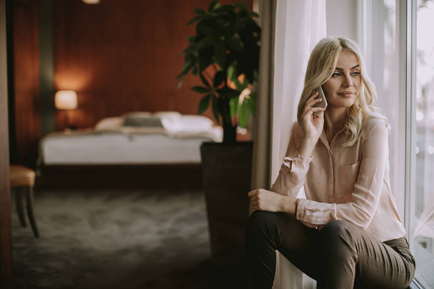 Pretty young blonde woman with mobile phone by the window in the room - Fotografie, Obrázek