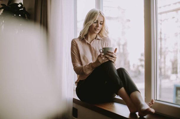 Blonde young woman relaxing and  drinking coffee by window in the room - 写真・画像