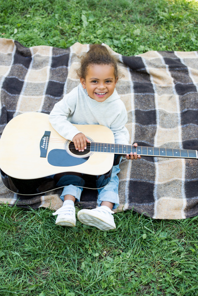 high angle view of smiling adorable african american kid holding acoustic guitar in park - Fotografie, Obrázek