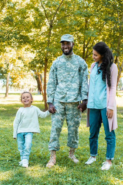 happy african american soldier in military uniform holding hands with daughter and wife in park - Photo, Image