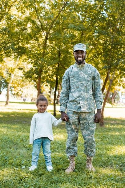 smiling african american soldier in military uniform and daughter holding hands in park - Fotografie, Obrázek