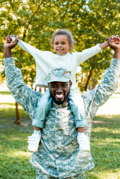 happy african american soldier in military uniform holding daughter on shoulders in park - Photo, Image