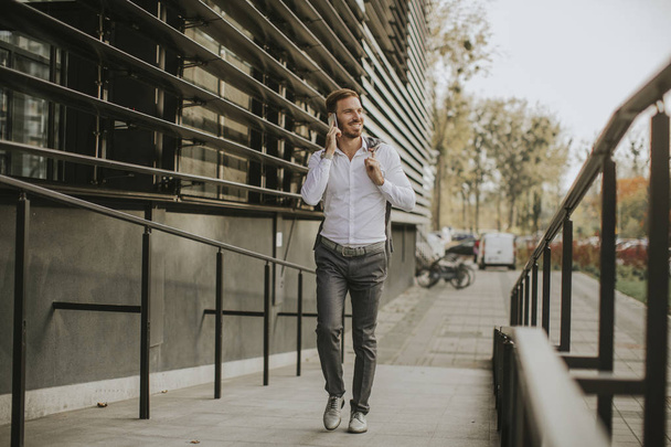 Knappe jonge zakenman wandelen met mobiele telefoon vóór kantoorgebouw - Foto, afbeelding