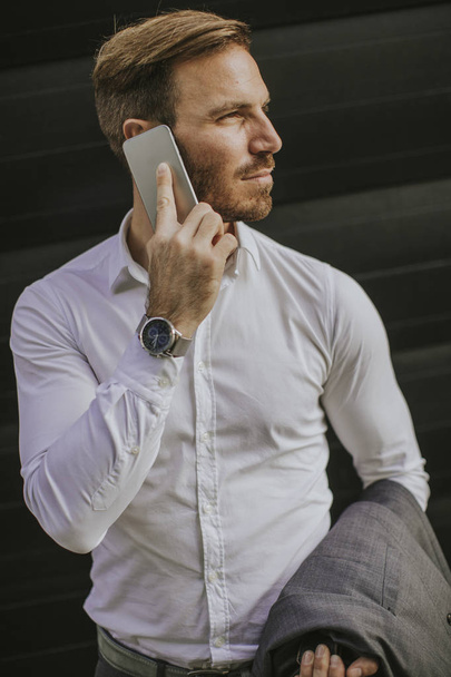 Joven empresario exitoso con traje gris y sosteniendo su teléfono inteligente mientras está de pie cerca de la oficina moderna o rascacielos
 - Foto, Imagen
