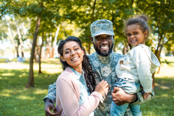 happy african american soldier in military uniform looking at camera with family in park - Photo, Image