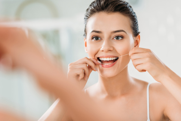 selective focus of beautiful smiling girl using dental floss in bathroom - Photo, Image