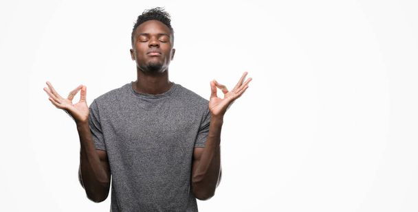 Jovem afro-americano vestindo camiseta cinza relaxar e sorrir com os olhos fechados fazendo gesto de meditação com os dedos. Conceito de ioga
. - Foto, Imagem