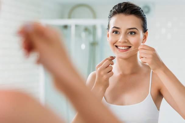 sorridente giovane donna in possesso di filo interdentale e guardando specchio in bagno
  - Foto, immagini