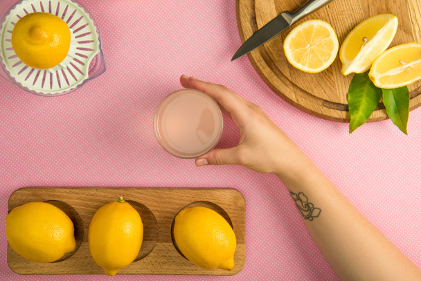top view of female hand with glass of lemon juice on pink table with squeezer and lemons on wooden boards - Photo, Image