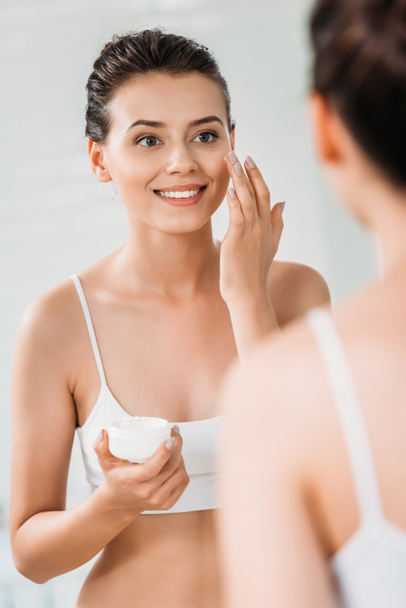 hermosa mujer joven sonriente aplicando crema facial y mirando el espejo en el baño
  - Foto, Imagen