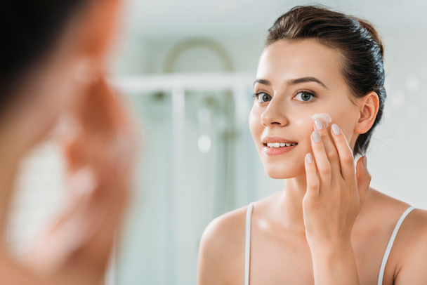 selective focus of smiling young woman applying face cream and looking at mirror in bathroom  - Foto, immagini