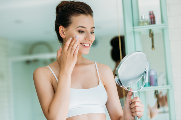 schöne lächelnde Mädchen mit Spiegel und Anwendung von Gesichtscreme im Badezimmer  - Foto, Bild