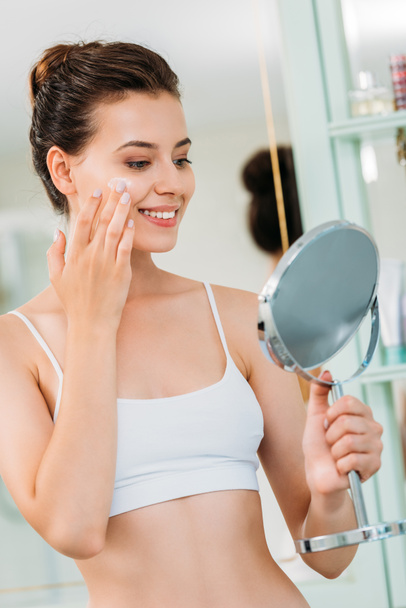 usměvavá mladá žena držící zrcadlo a použitím obličeje krémem v koupelně  - Fotografie, Obrázek