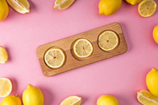 top view of fresh lemon slices on wooden board with lemons around on pink background - Foto, afbeelding