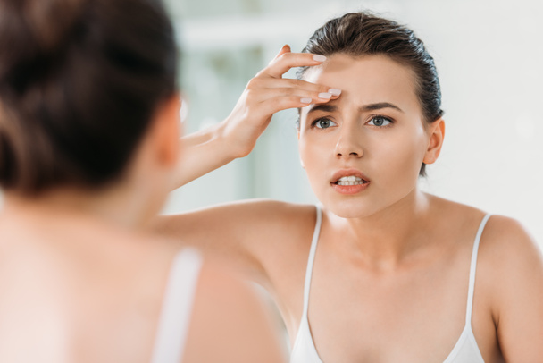attraktive junge Frau berührt Stirn und blickt im Badezimmer in den Spiegel - Foto, Bild