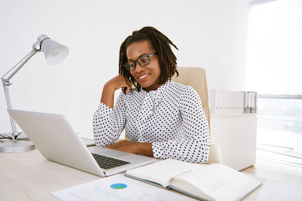 Africana dama de negocios mirando a la computadora portátil mientras está sentado en el escritorio con diario
  - Foto, Imagen