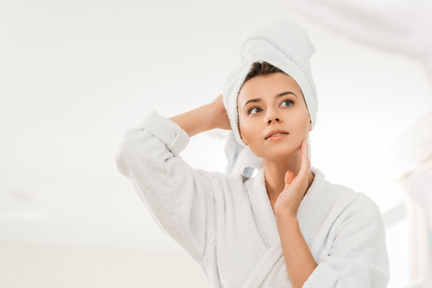 low angle view of beautiful pensive girl in bathrobe and towel on head looking away in bathroom   - Photo, Image