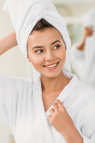 beautiful smiling girl in bathrobe and towel on head looking away in bathroom - Фото, изображение