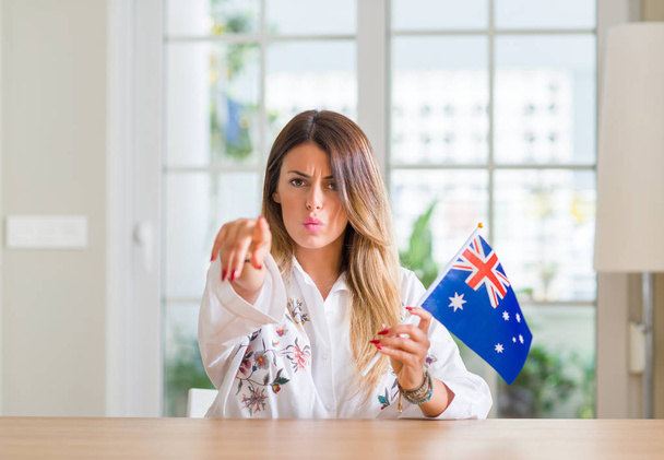 Jovem mulher em casa segurando bandeira da Austrália apontando com o dedo para a câmera e para você, sinal de mão, gesto positivo e confiante da frente
 - Foto, Imagem