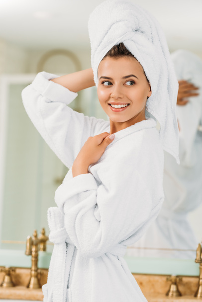 beautiful smiling girl in bathrobe and towel on head looking away in bathroom - Fotoğraf, Görsel