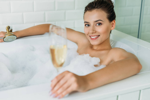 beautiful young woman holding glass of champagne and smiling at camera in bathroom - Foto, Imagen
